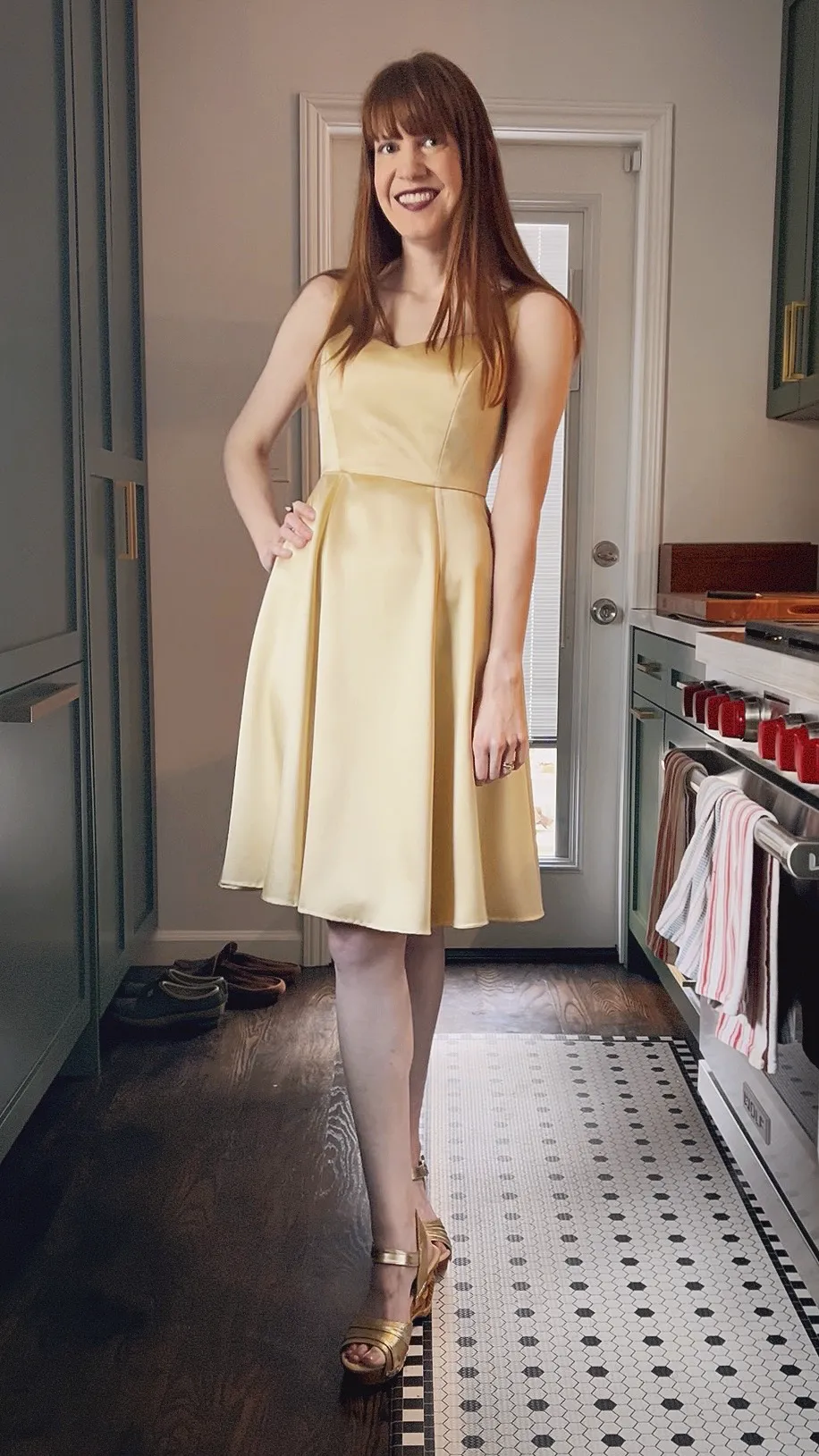 A woman standing in a kitchen, one hand on her hip. She is wearing a gold, knee-length silk dress with a sweetheart neckline and princess seams.